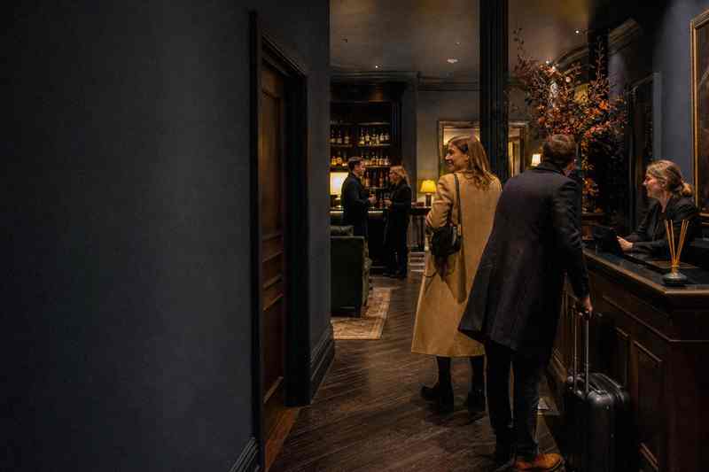 Een stel komt aan in de lobby van een klein hotel; de vrouw kijkt glimlachend om zich heen terwijl de receptioniste opkijkt